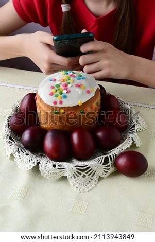 girl is sitting at table and taking pictures of traditional Easter cake on her phone. Red chicken eggs on white tablecloth. festive treat Easter.