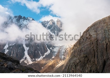 Scenic mountain landscape with snowy mountain top and glacier in dense low clouds in sunrise colors. Colorful mountain view to vertical glacier with icefall in morning sunlight in thick low clouds.