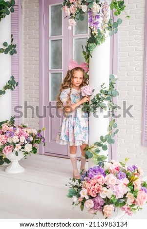 girl in a pink and blue dress stands on the veranda of a house with flowers. She has long blonde curls and a big pink bow on her head