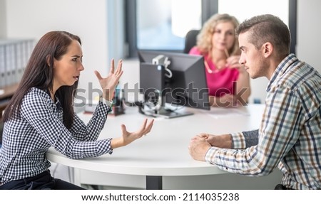 Woman gesticulates to a man angry over disagreement with demale mediator sitting behind the table in the background.