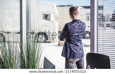 Trucking company manager looks out of the window and fleet of parked trucks during truck drivers shortage. Royalty-Free Stock Photo #2114035322