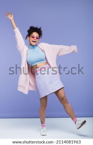 full length of cheerful african american woman with tattoo and outstretched hands on purple