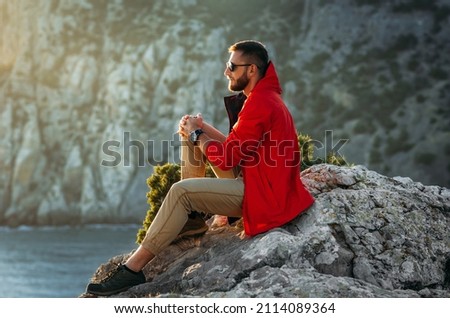 Portrait of a brutal bearded man in a red jacket by the sea. Male portrait on the background of a seascape. A traveler on the background of the sea coast. A man in a jacket at sunset Royalty-Free Stock Photo #2114089364