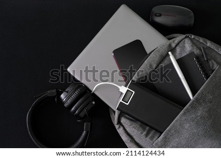 Silver laptop and modern gadgets lie in a gray backpack on a dark table. Travel, trip, study and lifestyle with modern computer technology. Close-up Royalty-Free Stock Photo #2114124434