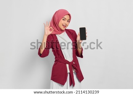 Cheerful beautiful Asian woman in casual shirt and hijab holding blank mobile phone screen, showing ok sign isolated over white background. People religious lifestyle concept Royalty-Free Stock Photo #2114130239
