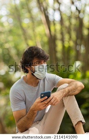 View of young man wearing a mask using a smartphone at day time with a green park in the background. Mobile phone, technology, urban concept. High quality photo