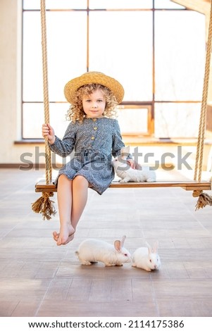 Girl child is cute and happy with white rabbits sitting on a rope swing at home against the background of a window in a braided hat