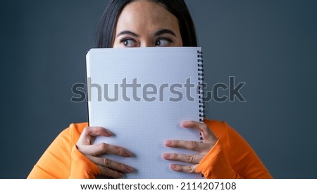 Young Asian Female Hiding Herself Behind a Big Notebook With Only Eyes Visible on a Grey Background With Copy Space on Left and Right, Wearing a Bright Orange Blouse.