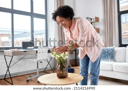 household, home improvement and interior concept - happy smiling young woman placing flowers on coffee table Royalty-Free Stock Photo #2114216990