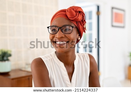 Cheerful mature black woman wearing spectacles and traditional turban at home. Beautiful african american woman with headscarf and eyeglasses sitting at home and looking away. Mature lady relaxing. Royalty-Free Stock Photo #2114224337