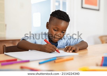 Little black boy sitting on table and painting with colored marker on book. Cute african american child drawing in the living room at home. Schoolboy colouring in book with red marker at home. Royalty-Free Stock Photo #2114224340