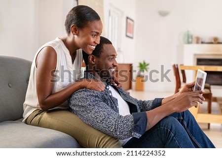 Middle aged black couple using digital tablet while sitting on couch to watch movie on streaming app. Happy black woman embracing boyfriend from behind while watching video on digital tablet at home. 