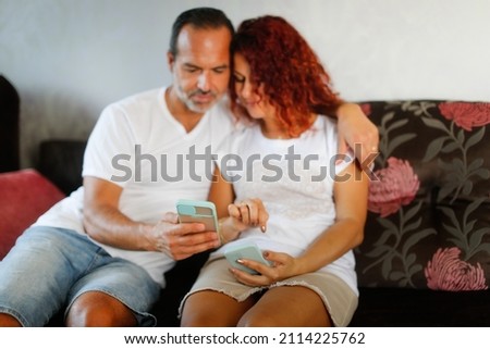 Cute middle-aged european couple with smart phones, cute middle-aged man and woman at home on the sofa show photos on the phone, search for information online on the internet. Help with gadgets
