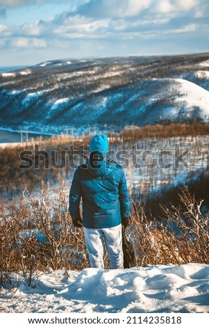  A young man stands on a high mountain with a beautiful winter snowy landscape on a sunny day