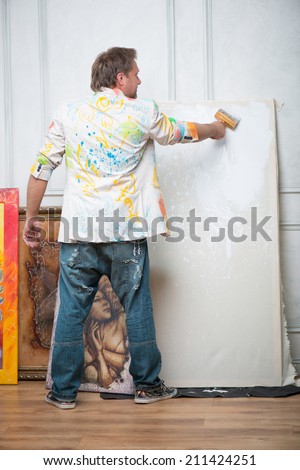 Full-length portrait of fair-haired painter wearing painted white shirt and blue torn jeans standing back to us among his pictures bleaching the linen