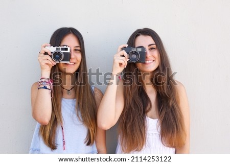 Two fashion girls with long hair on a white background take a picture with their retro cameras while smiling at the photographer. Best friends and emotion concept.