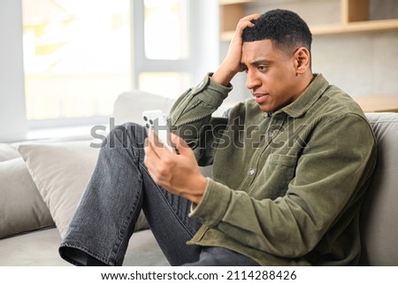 Sad young multiracial man holding smartphone and sitting on the sofa at modern apartment while looking with shocked face and holding his head Royalty-Free Stock Photo #2114288426