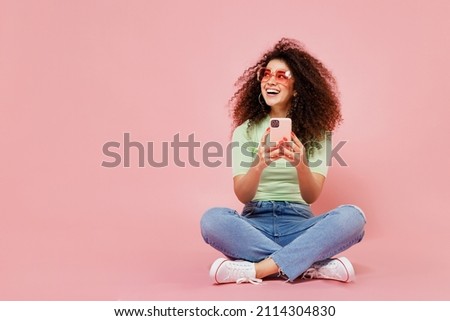 Full size young curly latin woman 20s wears casual clothes sit hold use mobile cell phone look aside on workspace area copy space mock up isolated on plain pastel light pink background studio portrait Royalty-Free Stock Photo #2114304830
