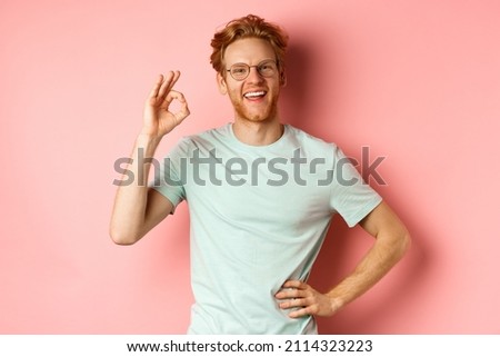 Cheerful guy with red hair and beard, wearing glasses, showing OK sign in approval and saying yes, smiling satisfied, standing over pink background