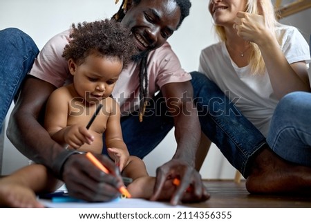 loving young bonding couple parents with daughter drawing pictures sitting on floor in living room, happy family weekend pastime. at home. black man, caucasian woman with child, domestic atmosphere