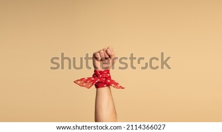 A man's hand, with a red bandana with white polka dots on his wrist, showing his fist. Feminist Movement. Beige background. Royalty-Free Stock Photo #2114366027