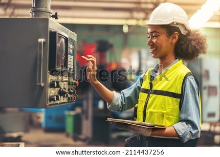 Standing in front of a control panel, a female industrial electrical engineer with a safety hardhat on her head and a tablet in her hand checks and maintains CNC machines in a factory.