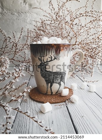 a ceramic mug with painted deer stands on a wooden table surrounded by branches with small white flowers. Breakfast with cocoa and marshmallows