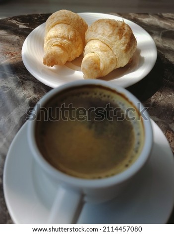 Closeup view vertical stock photography of white cup of hot black coffee and two fresh tasty croissants laying on white plate standing on brown table background. Morning breakfast concept