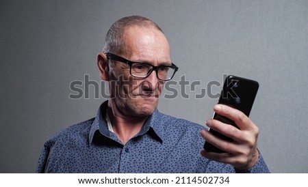 Senior man pensioner with bad eyesight in elegant black-rimmed glasses tries to read message in modern smartphone on grey background closeup. Royalty-Free Stock Photo #2114502734