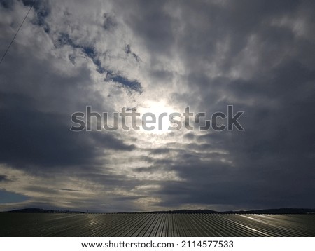 The shining sun through the gray accumulated clouds in the blue sky above the wide wavy reflective roof below and the black hilly line in the distance.