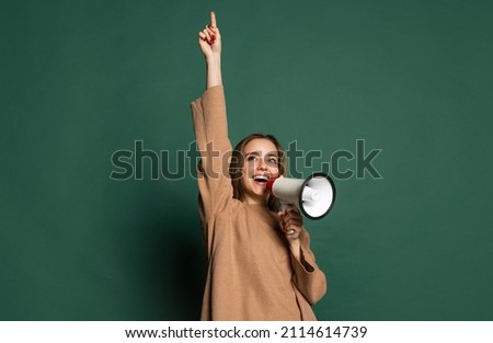 Attention. Portrait of young beautiful girl wearing brown sweatshirt shouting at megaphone isolated on green background. Concept of emotions, facial expression, youth, aspiration, sales.