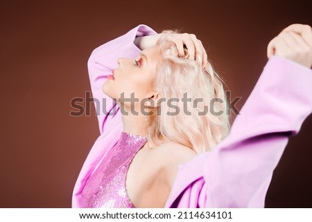 A blonde woman in a pink jacket, a jacket in sequins poses in the studio on a brown background. High fashion, 90s 80s. The girl dances and moves. Model with pink makeup