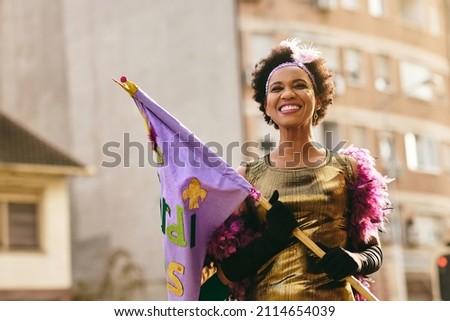 Happy African American woman holding Mardi Gras flag while wearing carnival costume and make-up on street parade. 