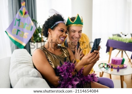 Happy African American woman and her male friend in Mardi Gras costumes using mobile phone at home party during the festival. 