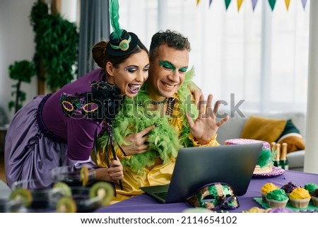 Happy couple greeting someone during video call over laptop while celebrating Mardi Gras festival at home.