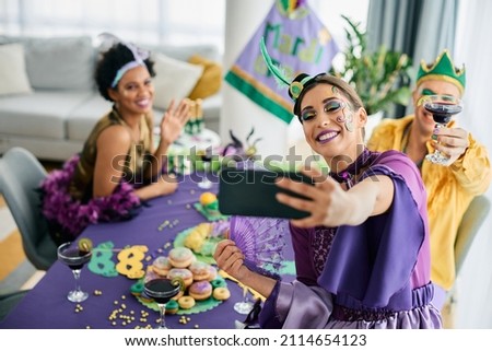 Group of cheerful friends in carnival costumes celebrating Mardi Gras and taking selfie with smart phone. 