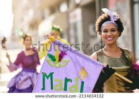 Happy African American woman in carnival costume holding Mardi Gras flag on street parade and looking at camera.