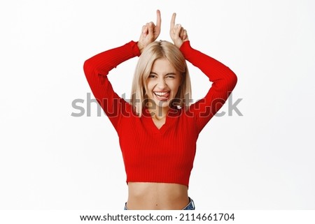 Coquettish and sassy attractive woman, showing finger horns, pointing fingers up, winking and sticking tongue silly, standing over white background