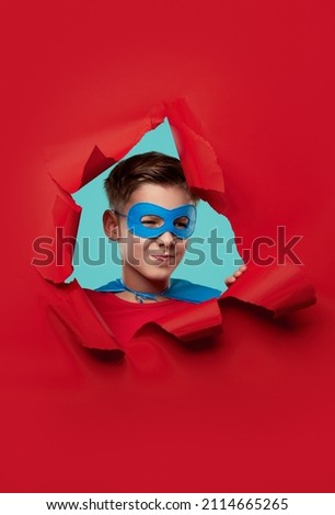 Adorable boy in superhero mask winking and looking at camera through red ripped paper background in studio