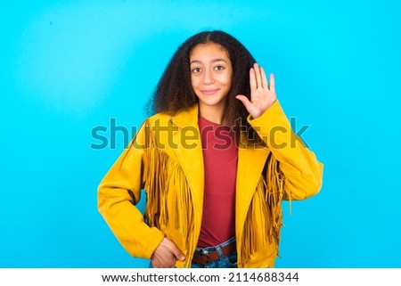 beautiful teenager girl wearing yellow jacket over blue background waiving saying hello or goodbye happy and smiling, friendly welcome gesture.