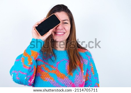 Young caucasian woman wearing vintage colorful sweater over white background holding modern smartphone covering one eye while smiling