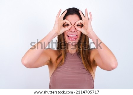 Young caucasian woman wearing tank top over white background doing ok gesture like binoculars sticking tongue out, eyes looking through fingers. Crazy expression.