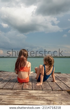 Small girls sitting on the lake wooden pier, enjoying summer vacation, talking about secrets, having fun. 