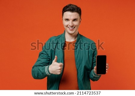 Young brunet man 20s wears red t-shirt green jacket hold in hand use mobile cell phone with blank screen workspace area show thumb up like gesture isolated on plain orange background studio portrait
