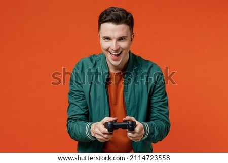 Gambling cheerful fun friendly fascinating happy young brunet man 20s wear red t-shirt green jacket hold in hand play pc game with joystick console isolated on plain orange background studio portrait. Royalty-Free Stock Photo #2114723558
