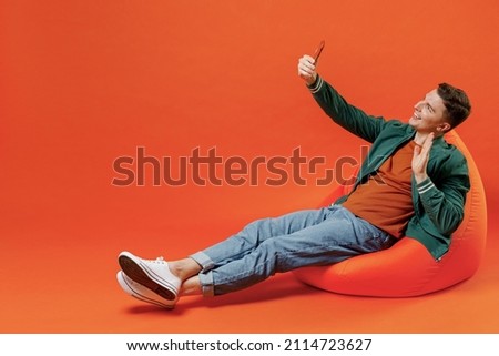 Full size body length young brunet man 20s wears red t-shirt green jacket sit in bag chair doing selfie shot on mobile cell phone greet waving hand isolated on plain orange background studio portrait. Royalty-Free Stock Photo #2114723627