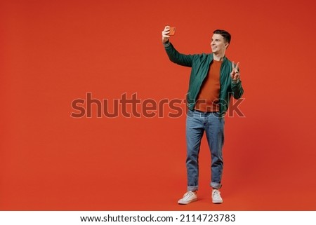 Full size body length happy smiling young brunet man 20s wears red t-shirt green jacket doing selfie shot on mobile cell phone showing victory sign isolated on plain orange background studio portrait