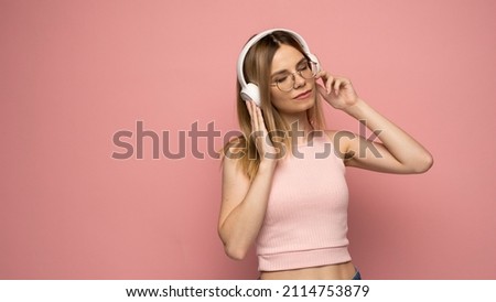 Pretty girl in a pink t-shirt and glasses listening music with her headphones and dancing on pink background.
