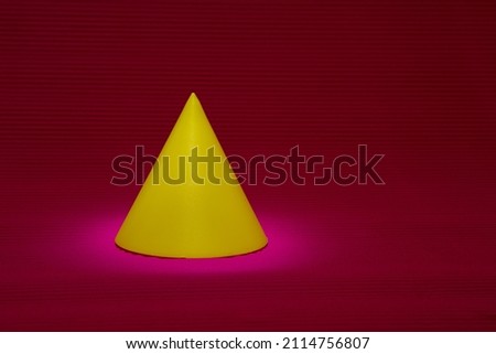 Conical geometric shape on serrated background in pink and red hues. Cone with yellow tint in selective focus. Yellow conical shape illuminated from above.
