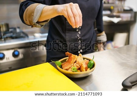 Unrecognizable cook sprinkling salt over a bowl of pork and hot peppers while working in the kitchen of a modern restaurant. Famous torreznos of Spanish cuisine Royalty-Free Stock Photo #2114771093